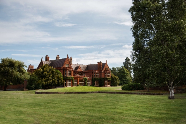 Holmewood Hall, panoramic view from lawn looking at red brick historic house with trees surrounding it