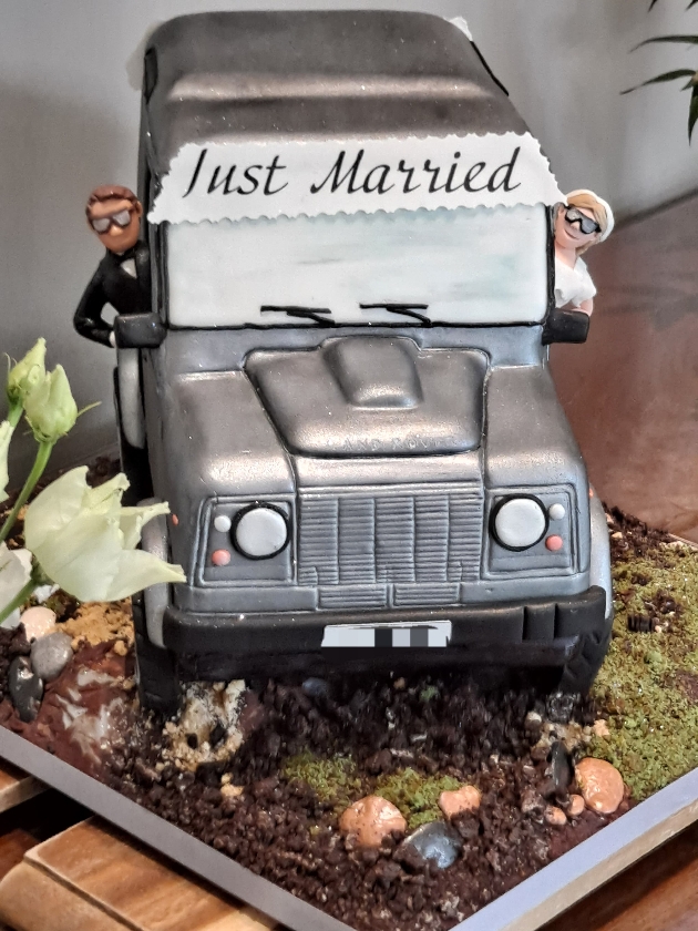 cake in the shape of a range rover with a bride and groom hanging out of it 