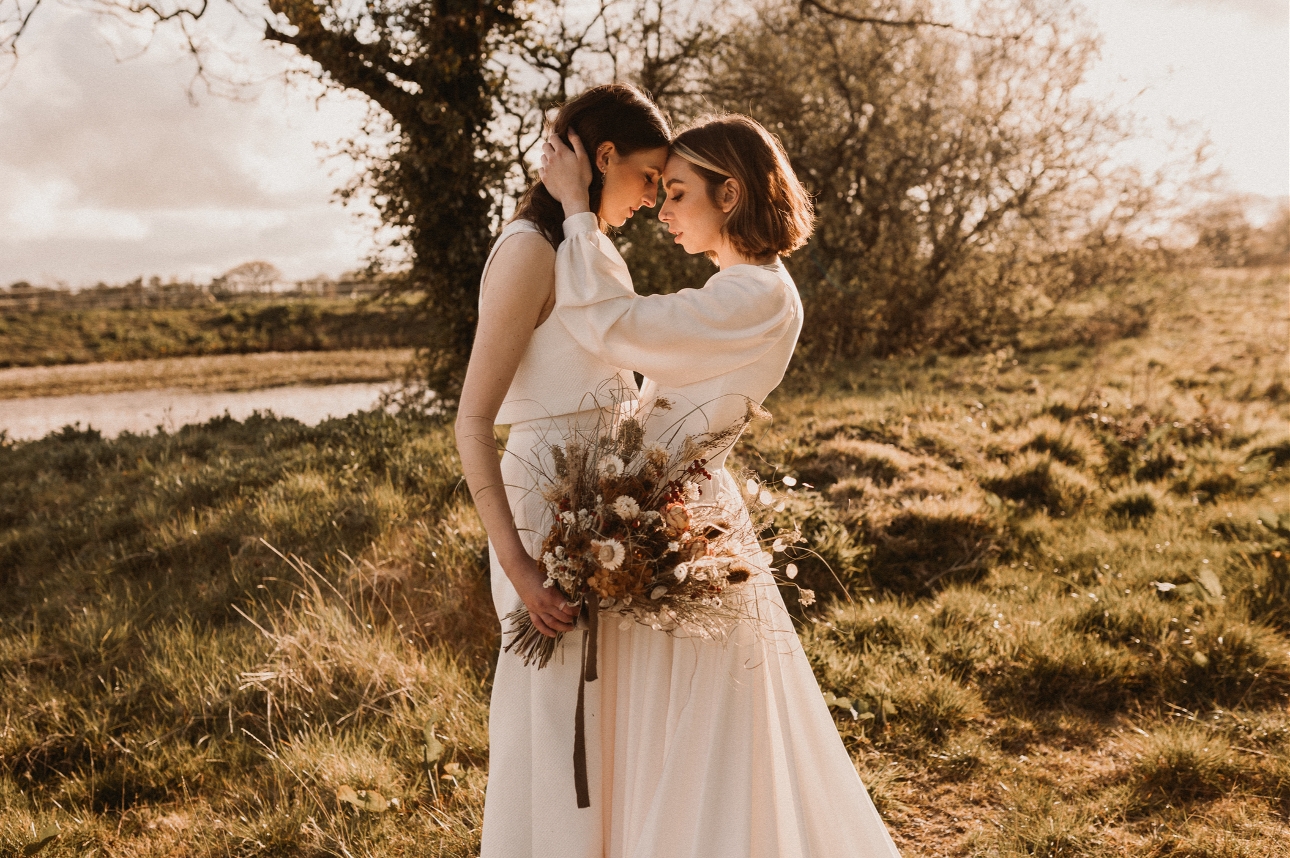 couple in wedding dresses head to head one holding a bouquet of boho flowers while standing a field