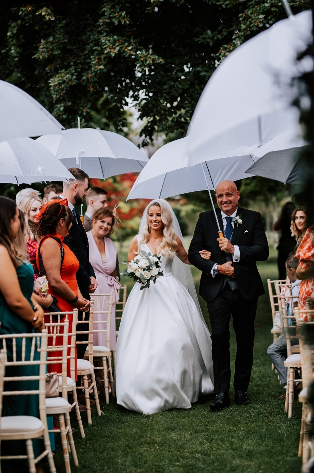 Father holds umbrella over bride as they walk down the aisle