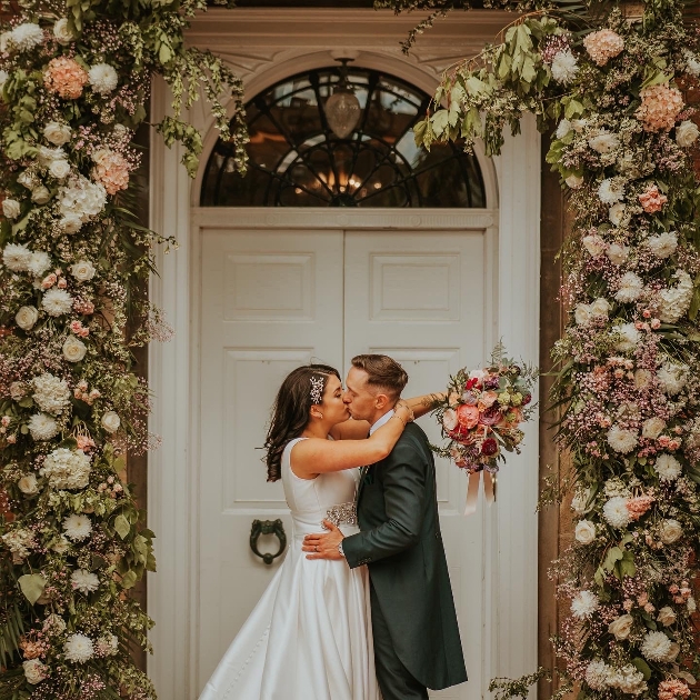 bride and groom in front of white door kissing and masses of flowers around in an arch