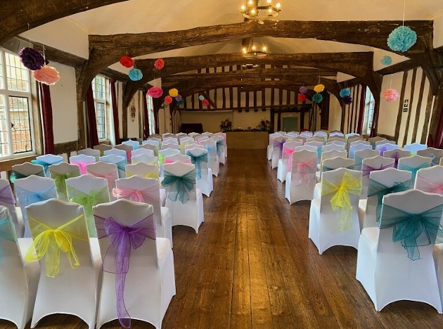 The inside of a barn with rows of white chairs with different coloured bows on the back of them