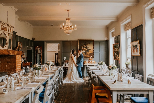 A bride and groom embracing in a large cream room surrounded by two long tables