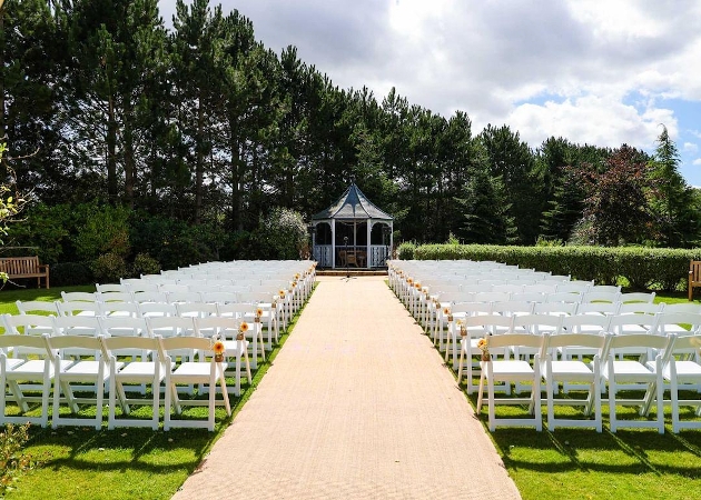 An outdoor ceremony set up with white chairs and a gazebo at the end of the aisle