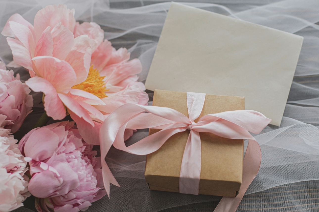 present wrapped in brown paper and tied in pink bow. Laying on organza next to peonies and a card