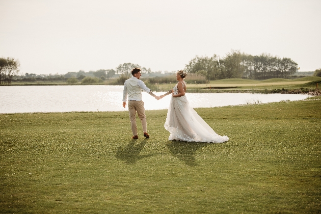 A bride and groom holding hands while walking on grass towards a pond